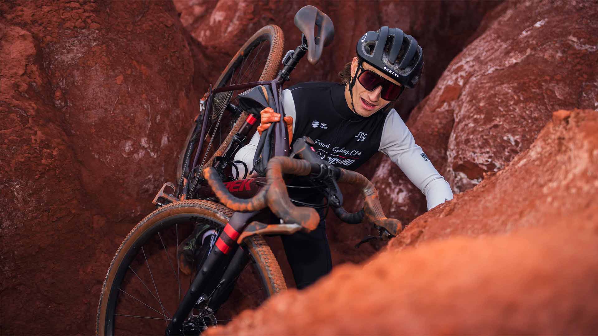 Athlète en plein effort portant son vélo TREK sur terrain escarpé rouge – photographie sport STUDIO M2X